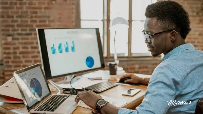 Homem negro de óculos analisando gráficos financeiros em monitor e notebook em escritório de tijolos à vista.