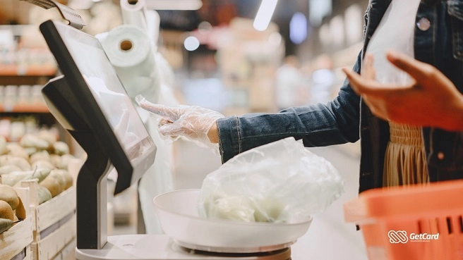 Cliente de luvas usando tela de autoatendimento para pesar frutas em supermercado moderno com foco no terminal.