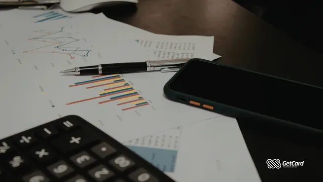 Calculadora, caneta e smartphone sobre papéis com gráficos financeiros coloridos em mesa de escritório.