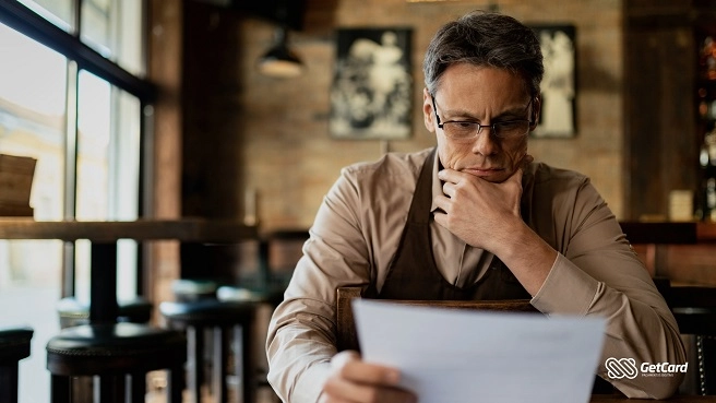 Homem pensativo com a mão no queixo lê um relatório impresso dentro de um café ou restaurante.