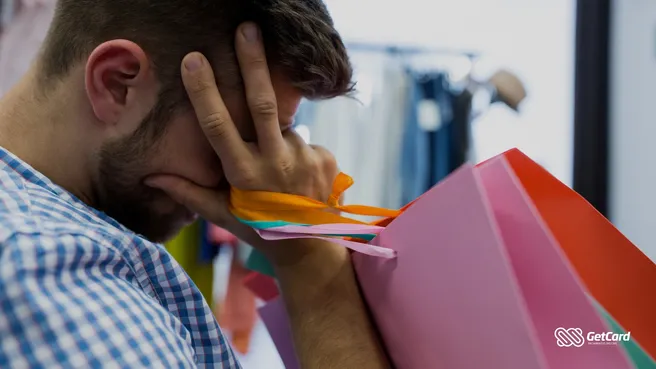 Homem jovem com as mãos no rosto em sinal de preocupação, segurando diversas sacolas de compras coloridas.