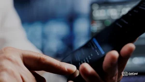 Close de mãos usando aplicativo de gestão financeira em smartphone com tela de monitor desfocada ao fundo.