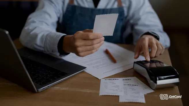 Empreendedor em uma mesa com laptop e máquina de cartão, analisando comprovantes de vendas e documentos financeiros.