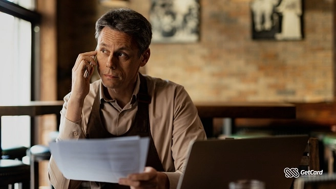 Homem fala ao celular enquanto segura documentos e olha para o lado, sentado em frente a um notebook em um café.