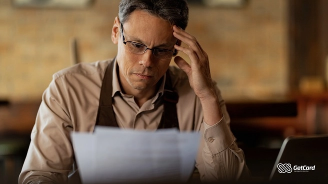 Homem de óculos com expressão preocupada segura papéis e analisa contas em um ambiente de escritório.