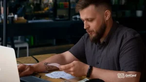 Homem jovem com barba e tatuagem analisa recibos de papel em uma mesa de madeira com um notebook aberto.