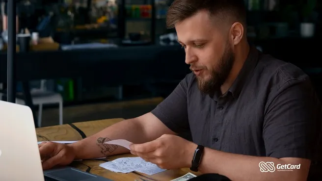 Homem jovem com barba e tatuagem analisa recibos de papel em uma mesa de madeira com um notebook aberto.