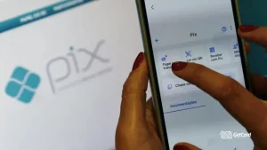 Mãos de mulher usando smartphone para realizar pagamento via Pix com o logotipo do Pix ao fundo.