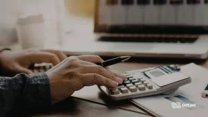 Mão de pessoa usando calculadora e caneta sobre mesa com notebook e gráficos de gestão. Logotipo GetCard no canto inferior.