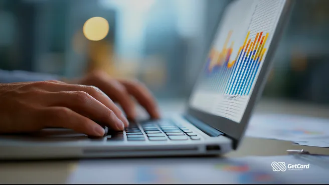 Mãos digitando em notebook com gráficos financeiros coloridos na tela e papéis de gestão sobre mesa de escritório.