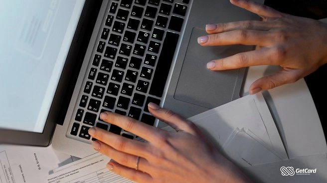 Vista superior de mãos femininas digitando em teclado de notebook prata sobre mesa com documentos de escritório.